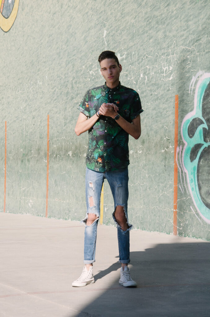Victor Zepeda stands against a graffiti-covered wall wearing a tropical-patterned shirt distressed jeans and white sneakers.